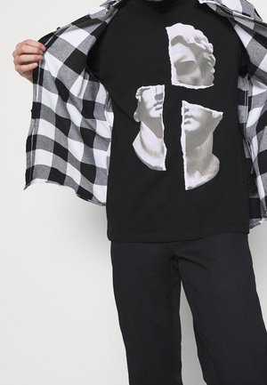 Osoba ubrana w czarne spodnie i czarną koszulkę z nadrukiem podartych białych klasycznych rzeźb, trzyma otwartą czarno-białą koszulę w kratkę.