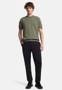 Olivgrünes Baumwoll-T-Shirt mit kurzen Ärmeln, kombiniert mit schwarzer Hose und schwarzen Schnürschuhen, das eine legere, moderne Passform zeigt.