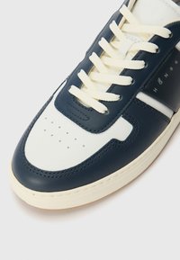 Σκούρο μπλε δερμάτινο αθλητικό παπούτσι με λευκές λεπτομέρειες, υφή στην επάνω πλευρά, στρογγυλή μύτη, τρυπήματα για αναπνοή και επίπεδα κρεμ κορδόνια.