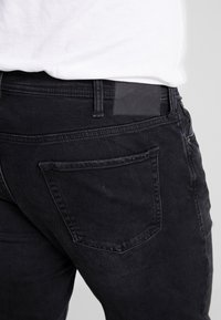 Vaqueros de mezclilla negra con corte ajustado. Incluye bolsillos traseros con costuras decorativas y un parche de cuero negro liso en la cinturilla.