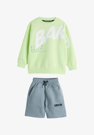Garçon souriant debout, portant un sweat-shirt vert clair avec un grand texte blanc, un short gris, des chaussettes blanches et des chaussures blanches sur un sol gris.