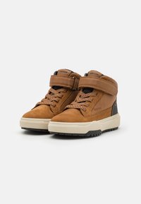Bottines en cuir marron avec suède texturé et semelle en caoutchouc. Elles possèdent une sangle Velcro et des lacets. Semelle extérieure antidérapante avec des accents noirs.