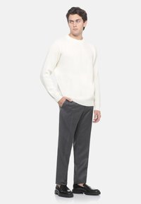 Maglione bianco a coste con maniche lunghe, abbinato a pantaloni grigi su misura e scarpe nere slip-on. Il modello posa con le mani in tasca.