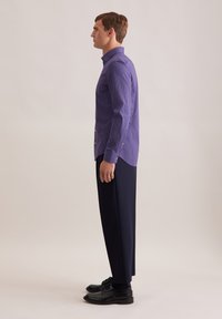 Camicia viola a maniche lunghe con un motivo geometrico, abbinata a pantaloni navy e scarpe nere. La camicia presenta un colletto con bottoni e polsini.