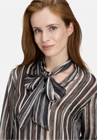 Vrouw met roodharen die een gestreepte blouse draagt met een grote strik om de hals, die vriendelijk naar de camera lacht.