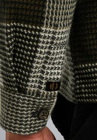 Chemise en tissu tissé dans un motif vichy gris et vert, avec un bouton noir et une étiquette de marque PME Legend sur la manche.