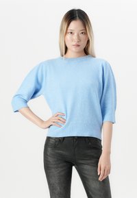 Mlada ženska z ravnimi blond lasmi, oblečena v svetlo modro pulover s tremičetrtnimi rokavi in črnimi strukturiranimi hlačami, stoji z roko na boku.