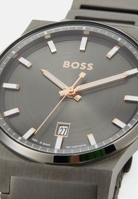 Reloj de hombre BOSS con caja metálica gris, esfera minimalista, acentos en oro rosa, marcadores de hora blancos y una ventana de fecha a las 27. Correa de goma negra.