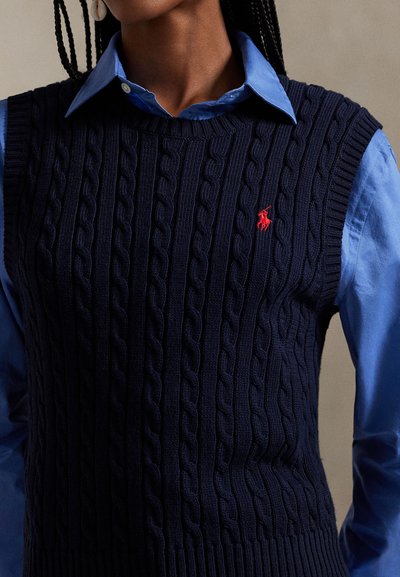 Γιλέκο πουλόβερ σε ναυτικό μπλε με πλεκτό σχέδιο, με ριμπ άκρες και κόκκινο κεντητό λογότυπο. Φορεμένο πάνω από ένα γαλάζιο πουκάμισο με γιακά.