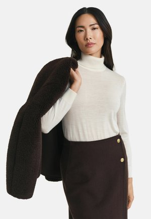 Frau mit langen dunklen Haaren, die einen weißen Rollkragenpullover und einen dunkelbraunen, hochgeschnittenen Rock trägt, hält einen dunkelbraunen, strukturierten Mantel über ihrer Schulter.