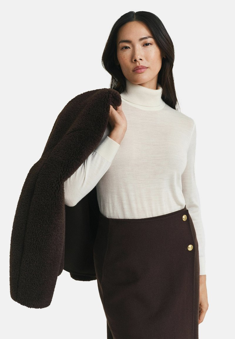 Frau mit langen dunklen Haaren, die einen weißen Rollkragenpullover und einen dunkelbraunen, hochgeschnittenen Rock trägt, hält einen dunkelbraunen, strukturierten Mantel über ihrer Schulter.