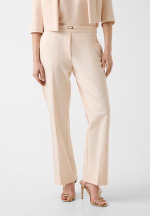 Femme portant un pantalon beige sur mesure et une veste courte assortie avec des sandales à talons hauts dorées à brides, debout sur un sol blanc.