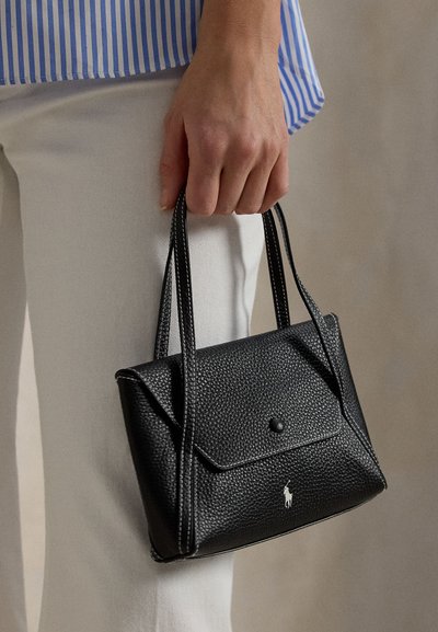 Χέρι που κρατάει μια μικρή μαύρη υφασμάτινη δερμάτινη τσάντα με διπλές λαβές και μπροστινό καπάκι, φορεμένη με λευκό παντελόνι και μπλε ριγωτό πουκάμισο.