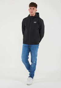 Juoda užsegama striukė su gaubtu, pagaminta iš lengvos medžiagos; krūtinėje yra logotipas, derinama su mėlynais džinsais ir baltais sportbačiais.