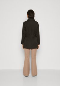 Trench coat nero con cintura, maniche lunghe, colletto e taglio dritto, abbinato a pantaloni beige chiaro a zampa su uno sfondo neutro.