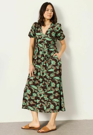 Woman wearing a brown midi dress with green and pink floral patterns, puff sleeves, V-neck, and tan slide sandals, standing against white wall.