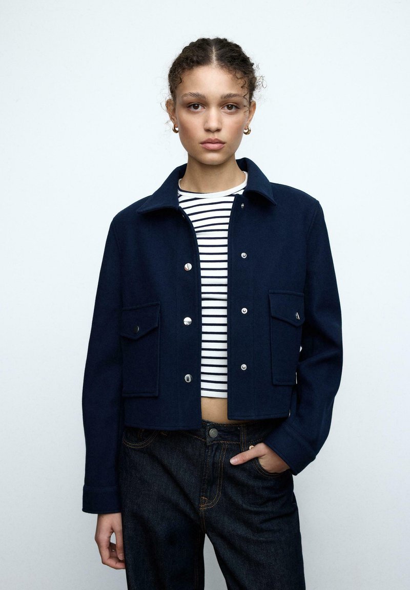 Ung kvinde iført en marineblå jakke med knapper over en sort-hvid stribet crop top og mørke jeans, stående med den ene hånd i lommen.