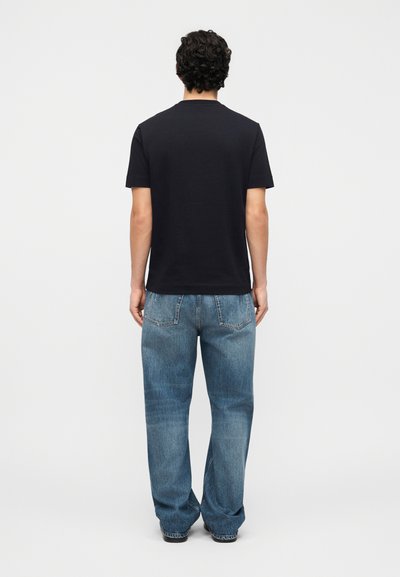 Άνδρας που στέκεται με την πλάτη προς τα εμπρός, φορώντας απλό μαύρο μπλουζάκι και φαρδιά μπλε τζιν, με φόντο λευκό.