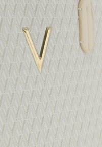 Borsa beige con motivo geometrico. Presenta un emblema dorato a forma di "V" e una superficie strutturata. Cuciture chiare evidenti su un dettaglio in pelle.