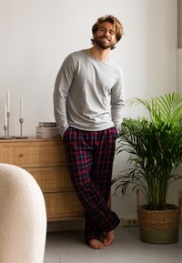 Grijze langemouwen shirt gecombineerd met rood en marineblauwe geruite flanellen broek. Staande bij een houten ladekast en een pot met een groene plant.