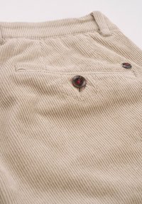 Beige Cordstoff mit vertikalen Rippen. Hat eine Gesäßtasche mit einem großen, dunklen Knopf und einem kleinen Logoakzent in der Nähe der Tasche.