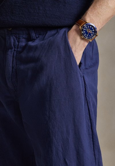 Nadgarstek z zegarkiem na brązowym skórzanym pasku i niebieską tarczą umieszczony jest w kieszeni luźnych granatowych spodni.