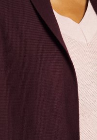 Gros plan d'une personne portant un blazer texturé violet foncé sur un pull en maille rose clair à col en V.