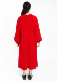 Červené šaty s dlouhým rukávem a vysokým límcem, volnými rukávy. Měkká, hladká tkanina splývá do poloviny lýtek. Jsou obuty černé robustní boty.