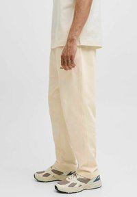 Cremefarvede løse sweatpants lavet af blødt stof, med sidesømme og et broderet logo nær lommen. Kombineret med grå og blå sneakers.