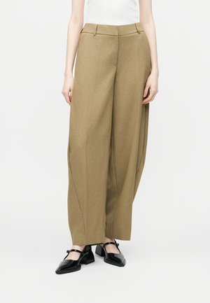 Kobieta ubrana w camelowe spodnie z wysokim stanem i szerokimi nogawkami z przodu z zakładkami oraz czarne buty Mary Jane, stojąca na białym tle.