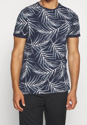 Hombre vestido con una camiseta azul marino con patrones de hojas de palmera en blanco y gris y pantalones negros, de pie con los brazos relajados a los lados.