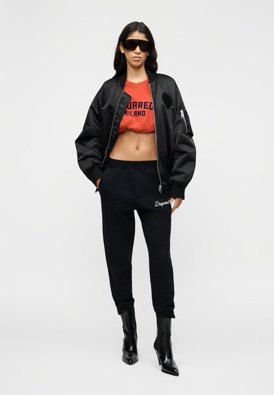 Veste bomber noire portée sur un t-shirt court rouge avec le texte "SQUARED MILANO". Pantalon de survêtement noir et bottes à talons hauts noires brillantes. Lunettes de soleil portées.