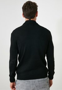 Pull noir en tricot avec un motif texturé, doté d'un col et de poignets côtelés. Le modèle porte un pantalon gris à carreaux.