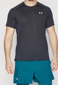 Muž ve černém krátkém rukávu sportovním tričku a tyrkysových šortsích s logy Under Armour na hrudi a na šortkách stojí proti jednoduchému pozadí.