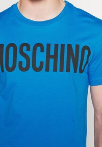 Detailní záběr na osobu v jasně modrém tričku s velkým černým nápisem "MOSCHINO" vytištěným přes hruď.