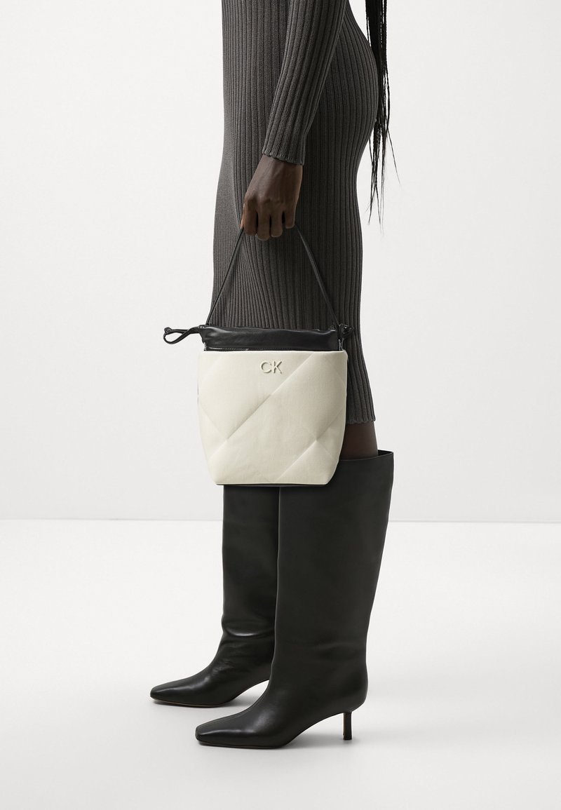 Personne en robe grise côtelée tenant un sac à main CK blanc et noir, portant des bottes hautes à talons noires, debout devant un fond uni.