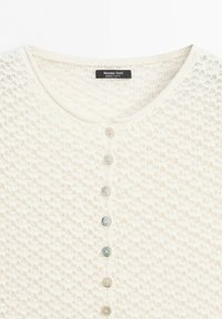 Close-up van een crèmegekleurde gebreide cardigan met een ronde halslijn en zes knopen in verschillende kleuren in het midden aan de voorkant.