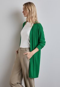 Cardigan vert en tissu doux, design à l'avant ouvert, manches longues, porté sur une chemise blanche et un pantalon beige, avec deux poches avant.
