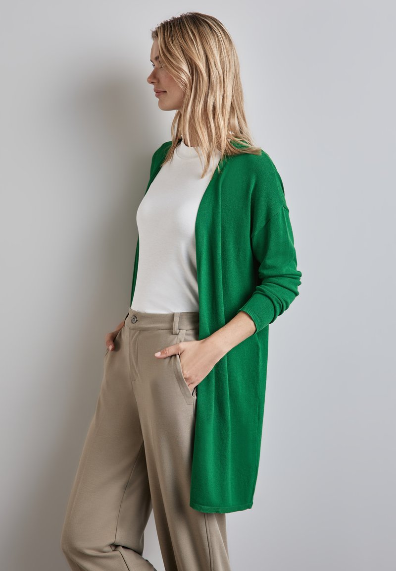 Cardigan vert en tissu doux, design à l'avant ouvert, manches longues, porté sur une chemise blanche et un pantalon beige, avec deux poches avant.