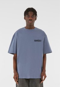 Tricou oversized albastru, din bumbac, cu mâneci scurte, guler rotund și o grafică neagră pe zona stângă a pieptului.