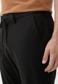 Zwarte broek met een zachte textuur, voorzien van een trekkoord in de taille en zijzakken, getoond dichtbij een hand die een bruine shirt draagt.