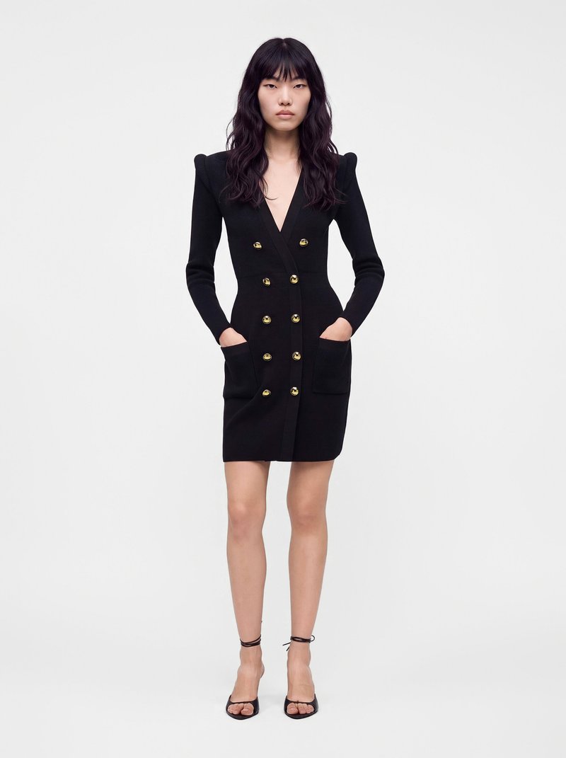 Femme portant une robe noire cintrée à double boutonnage avec des boutons dorés, manches longues, poches avant et talons ouverts noirs.