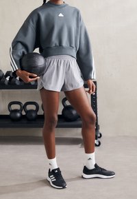 Grå kortärmad sweatshirt med vitt logotyp, ljusgrå shorts och svarta träningsskor. Modell som håller en svart medicinboll i en gymmiljö.