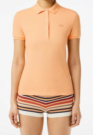 Frau trägt ein pfirsichfarbenes Poloshirt mit kurzen Ärmeln und mehrfarbige, gestreifte Strickshorts, steht vor einem weißen Hintergrund.