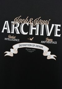 Crna majica s bijelim i smeđim tekstom. Sadrži natpise "Jack & Jones Archive", "Definition of Denim" te ukrasni dizajn vrpce.