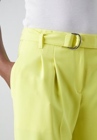Gros plan d’un pantalon jaune vif ceinturé avec des plis, porté avec un haut blanc côtelé, et une main reposant dans la poche.