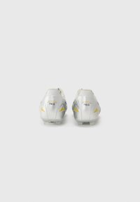 Crampons de football argentés avec une texture lisse, présentant des accents jaunes et un logo au niveau du talon. Conçus pour une adhérence sur les surfaces en herbe.