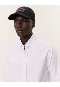 Camicia bianca con bottoni, caratterizzata da una texture sottile e un piccolo logo. Cappellino nero con testo ricamato "NIGHT SERVICE" in blu e rosso.