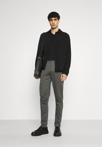 Veste noire à bouton sur une chemise noire, pantalon gris ajusté et baskets noires. Tient un sac en cuir noir, se tenant devant un fond blanc.