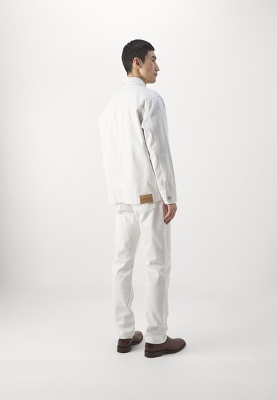 Veste et pantalon en denim blanc avec une coupe décontractée, arborant une étiquette en cuir brun, des détails de boutons et une texture lisse. Des chaussures brunes complètent le look.